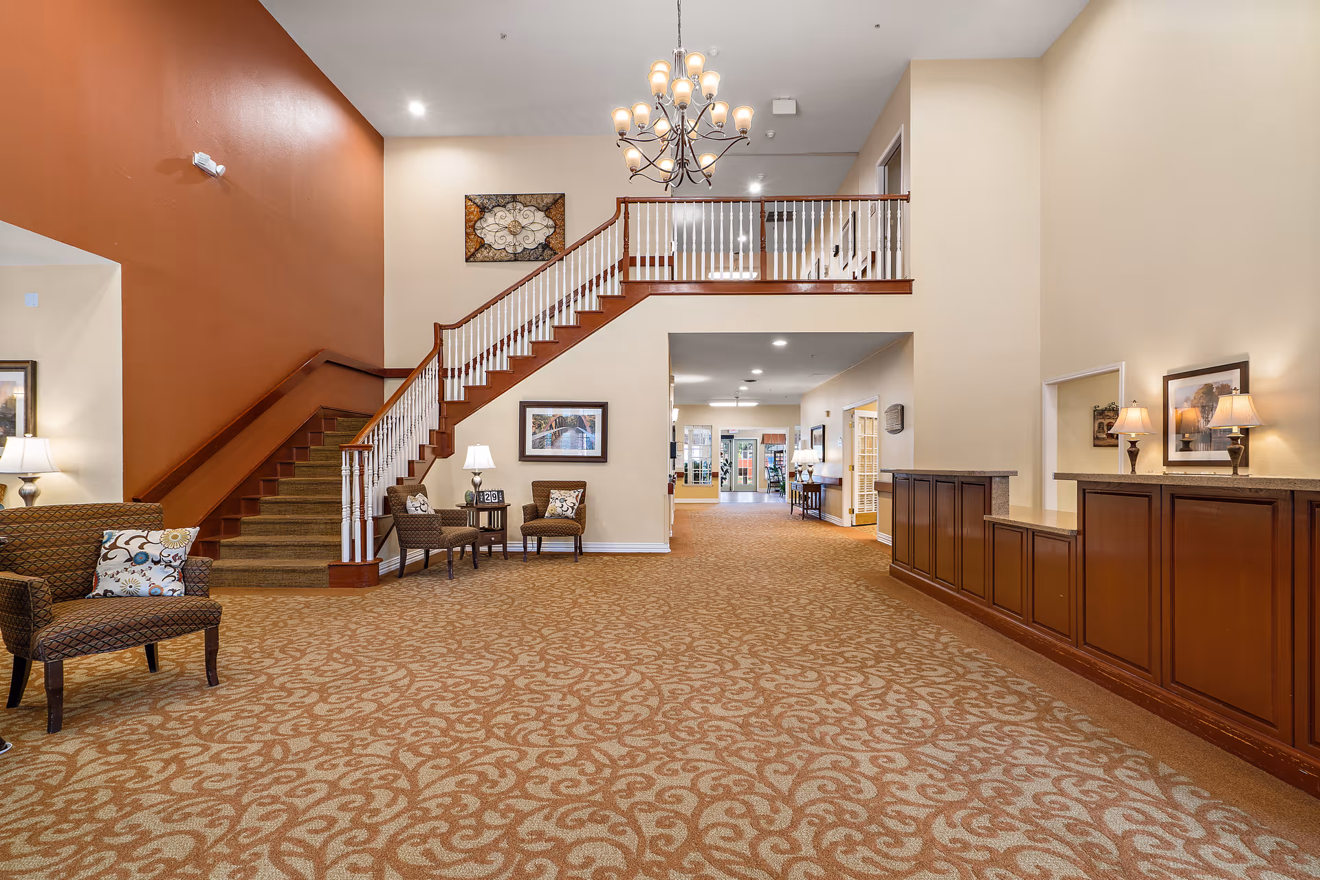 Spacious senior living facility lobby with patterned carpet, a staircase with wooden railing, several chairs with cushions, framed artwork on the walls, and a chandelier hanging from the ceiling. There is a reception desk on the right side with two table lamps and framed pictures above it.