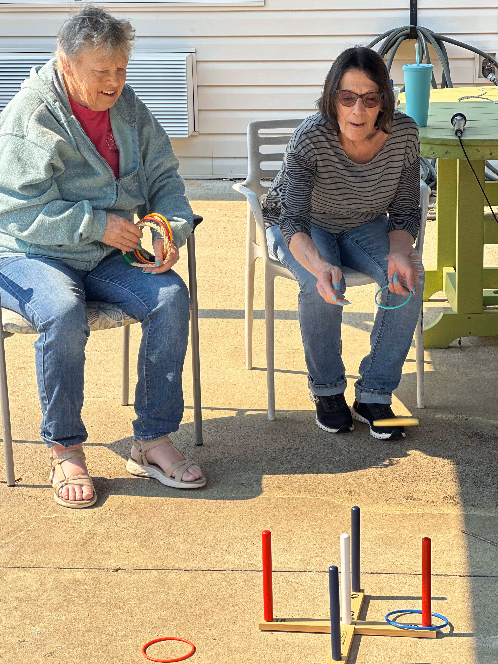 Two elderly women sitting outside on chairs playing a ring toss game on a concrete patio. One woman is holding several rings while the other is in the process of tossing a ring towards the game set. There is a green table with a blue cup and a hose reel in the background.