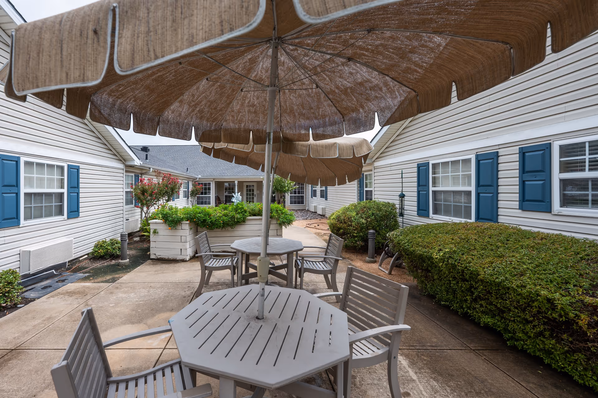 Outdoor courtyard area with beige patio umbrellas and gray tables and chairs. The space is surrounded by white buildings with blue shutters and has green bushes and plants along the walkway.