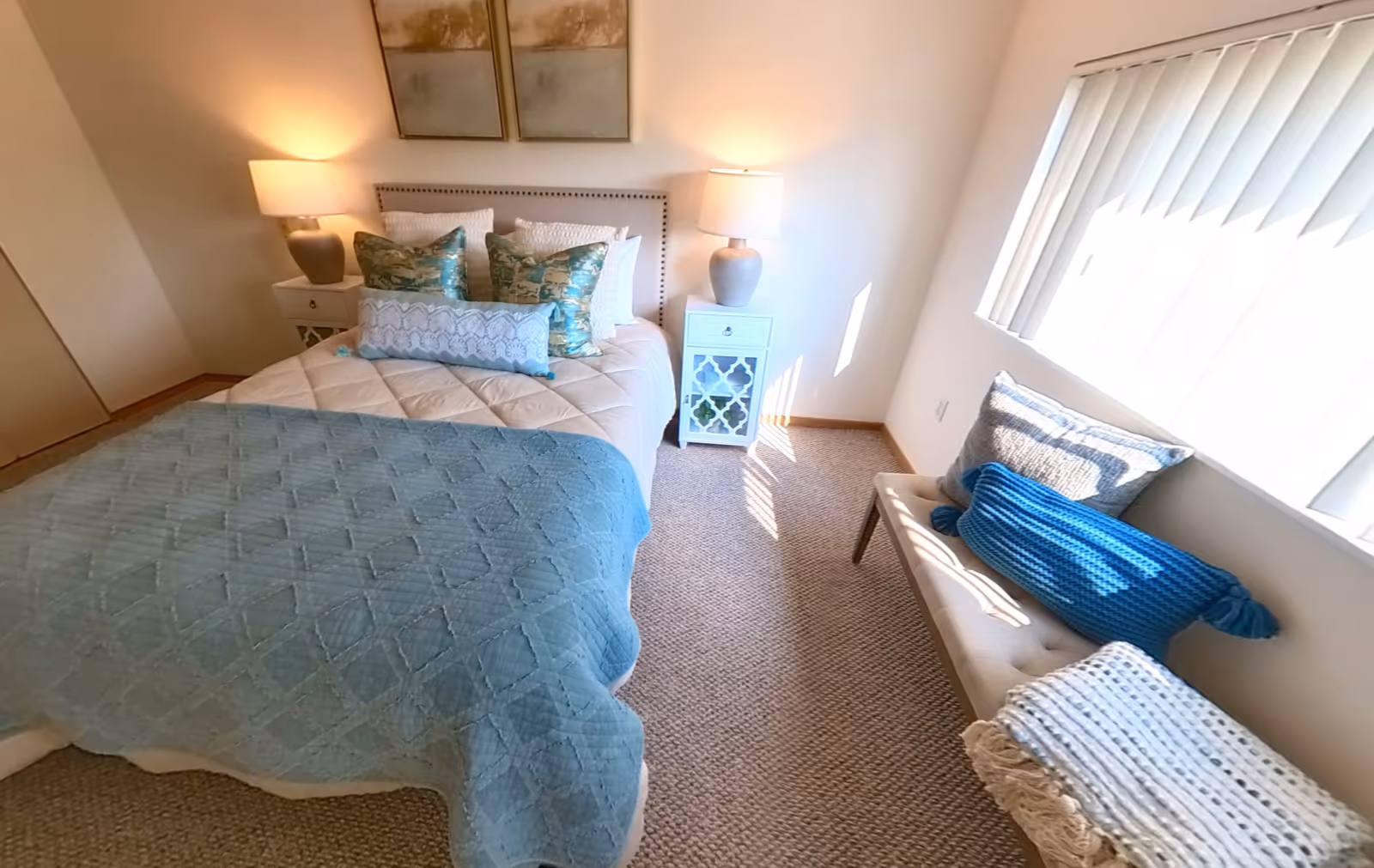 A cozy bedroom with a neatly made bed featuring a beige quilt and blue throw blanket. There are multiple decorative pillows on the bed. On either side of the bed are white nightstands with matching lamps. A cushioned bench with pillows and a folded blanket sits beneath a window with vertical blinds, allowing sunlight to stream in. The walls are light-colored, and the floor is carpeted.