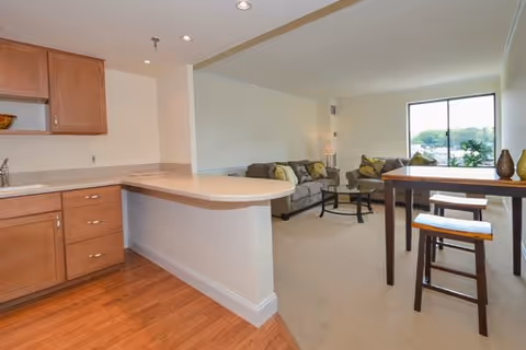 Interior view of a senior living apartment showing a kitchen area with wooden cabinets and a countertop that extends into a small breakfast bar. Adjacent to the kitchen is a living room with a sofa, coffee table, and a large window letting in natural light. There is also a tall dining table with two stools near the window.