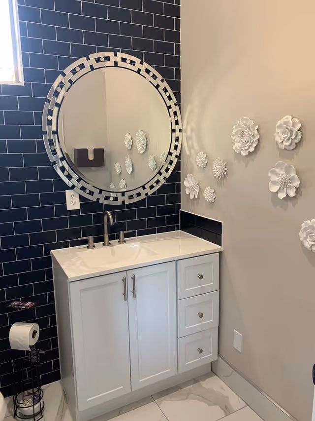 A modern bathroom vanity with a white cabinet and countertop, a silver faucet, and a large round mirror with a decorative frame mounted on a black tiled wall. The adjacent wall is beige and decorated with white 3D floral wall art. A toilet paper holder with a roll is visible on the left side.