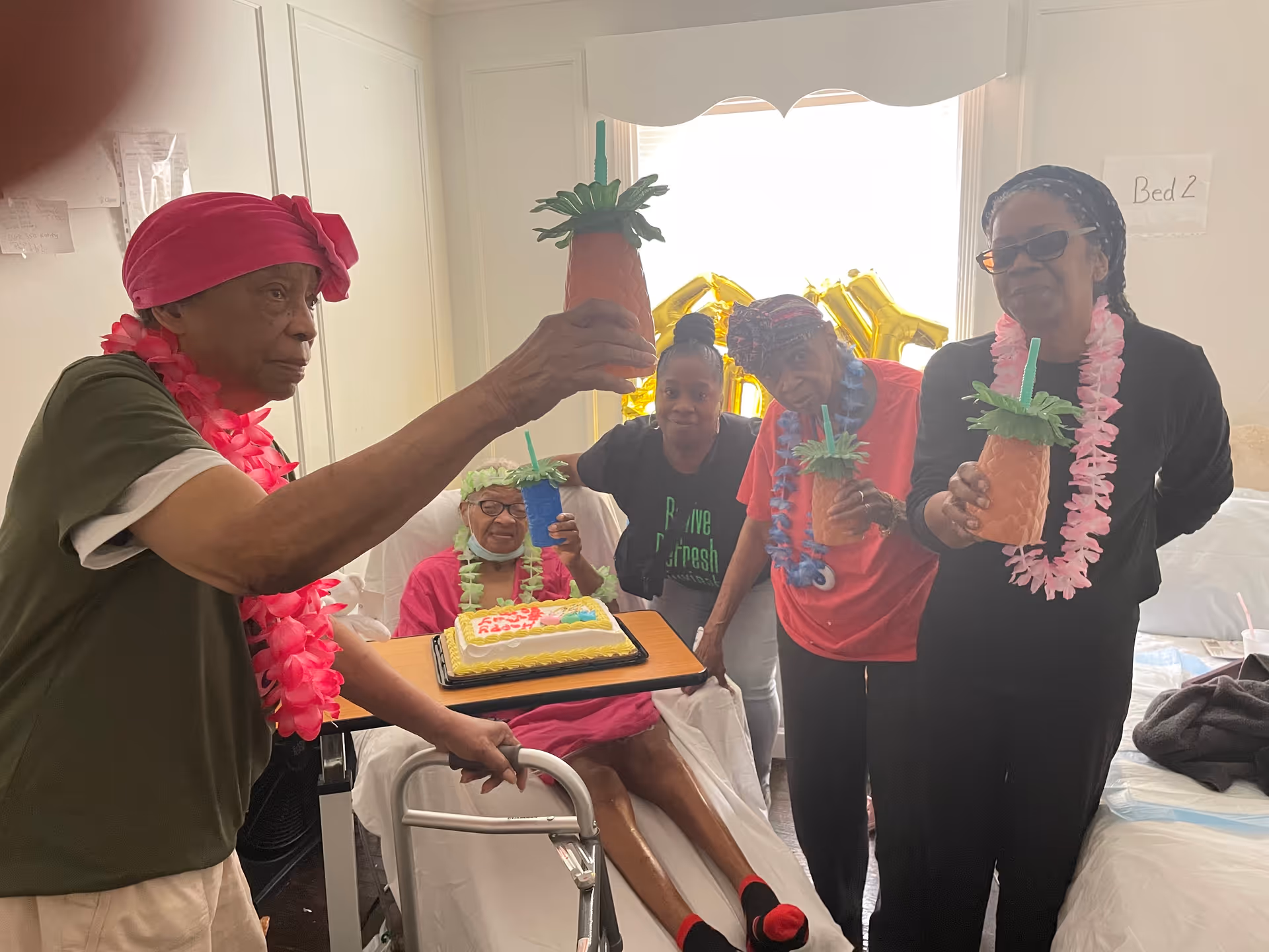 A group of five elderly women celebrating in a bedroom. One woman is sitting on a bed with a birthday cake on a tray, while the others stand around her holding pineapple-shaped cups with straws. They are wearing colorful leis and headscarves, and there are gold balloon letters in the background near a window.