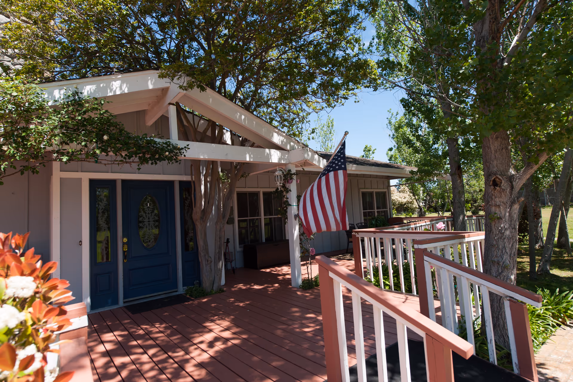 Front exterior view of a single-story building with a blue door and an American flag mounted on the porch. The porch has a wooden ramp with white railings, surrounded by trees and greenery under a clear blue sky.