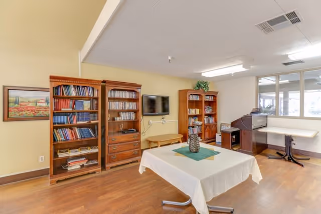 A bright common room with bookshelves, a wall-mounted TV, and a table covered with a white cloth in the center.