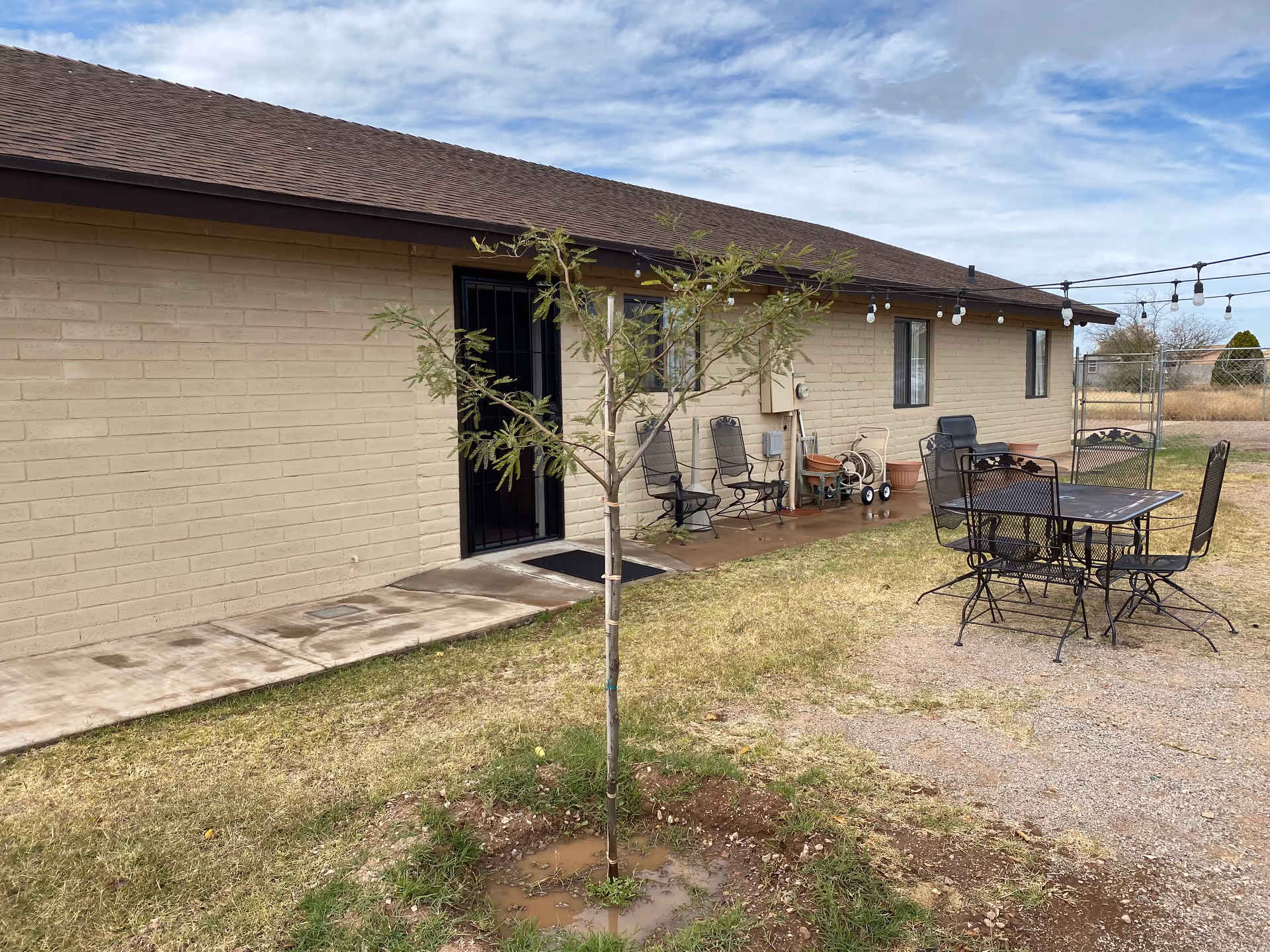 Outdoor patio area at Lesan Assisted Living featuring a small tree planted in the grass, a metal table with four chairs on a gravel surface, and a beige brick building with a brown roof in the background. There are string lights hanging above the patio and several chairs lined up against the building wall.