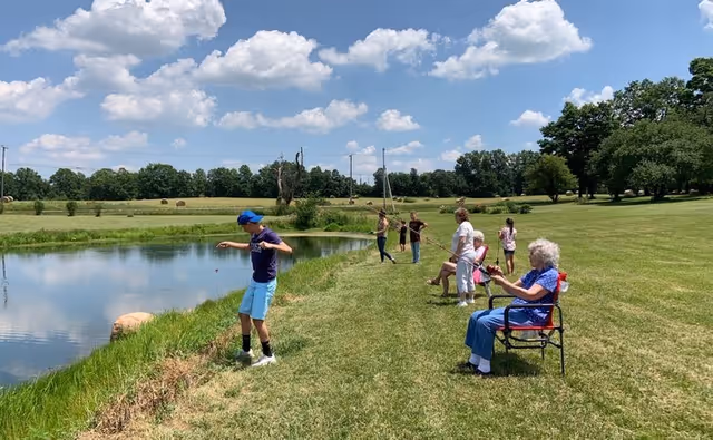 A group of people fishing by a pond on a sunny day with a partly cloudy sky. Several individuals are standing near the water's edge holding fishing rods, while two elderly women are seated on chairs on the grassy bank. The background features open fields, trees, and power lines.