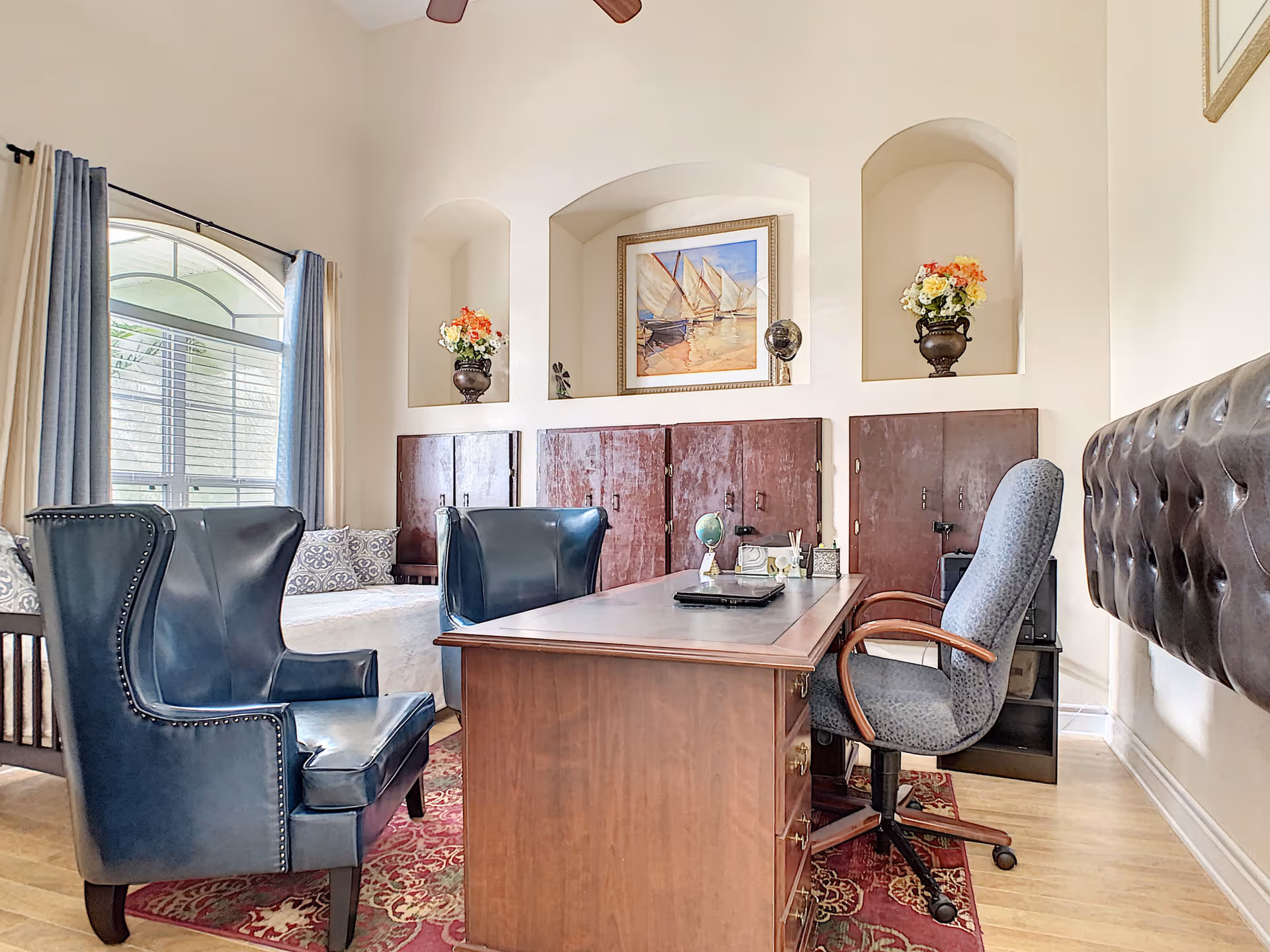 A cozy office or study area with a wooden desk, a gray office chair, and two blue leather armchairs. Behind the desk is a wall with three recessed niches, two holding flower vases and the center one displaying a framed painting of sailboats. A window with blue and beige curtains lets in natural light, and a patterned red rug covers the floor.