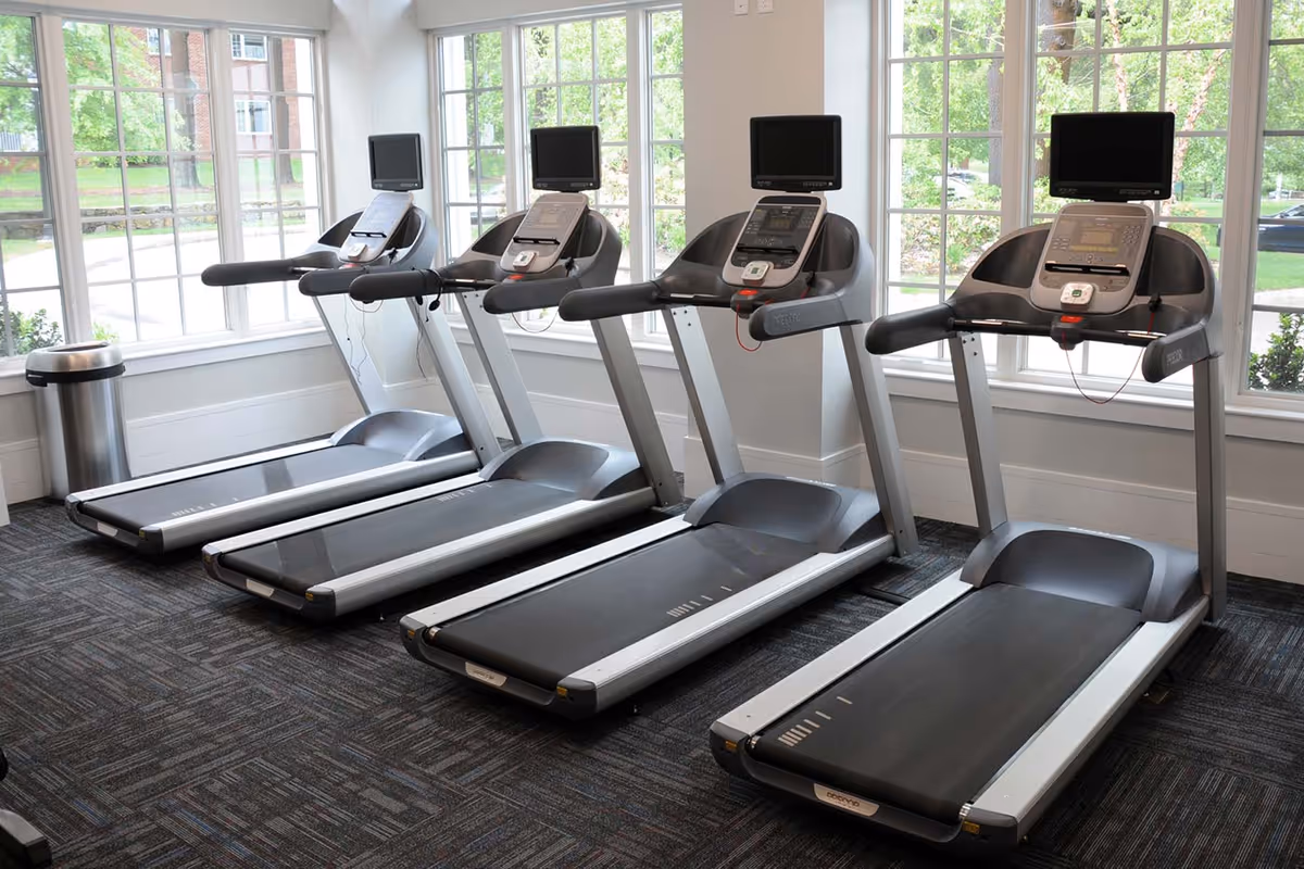 A fitness room with four treadmills lined up side by side in front of large windows that let in natural light. Each treadmill has a screen for workout tracking. A trash can is visible near the windows.