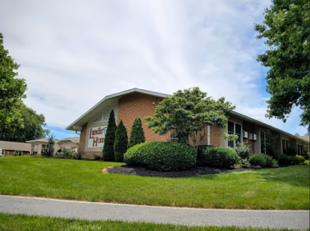 Exterior view of a single-story brick building with a sign that reads 'Landis Homes'. The building is surrounded by well-maintained green grass, bushes, and trees under a partly cloudy sky.