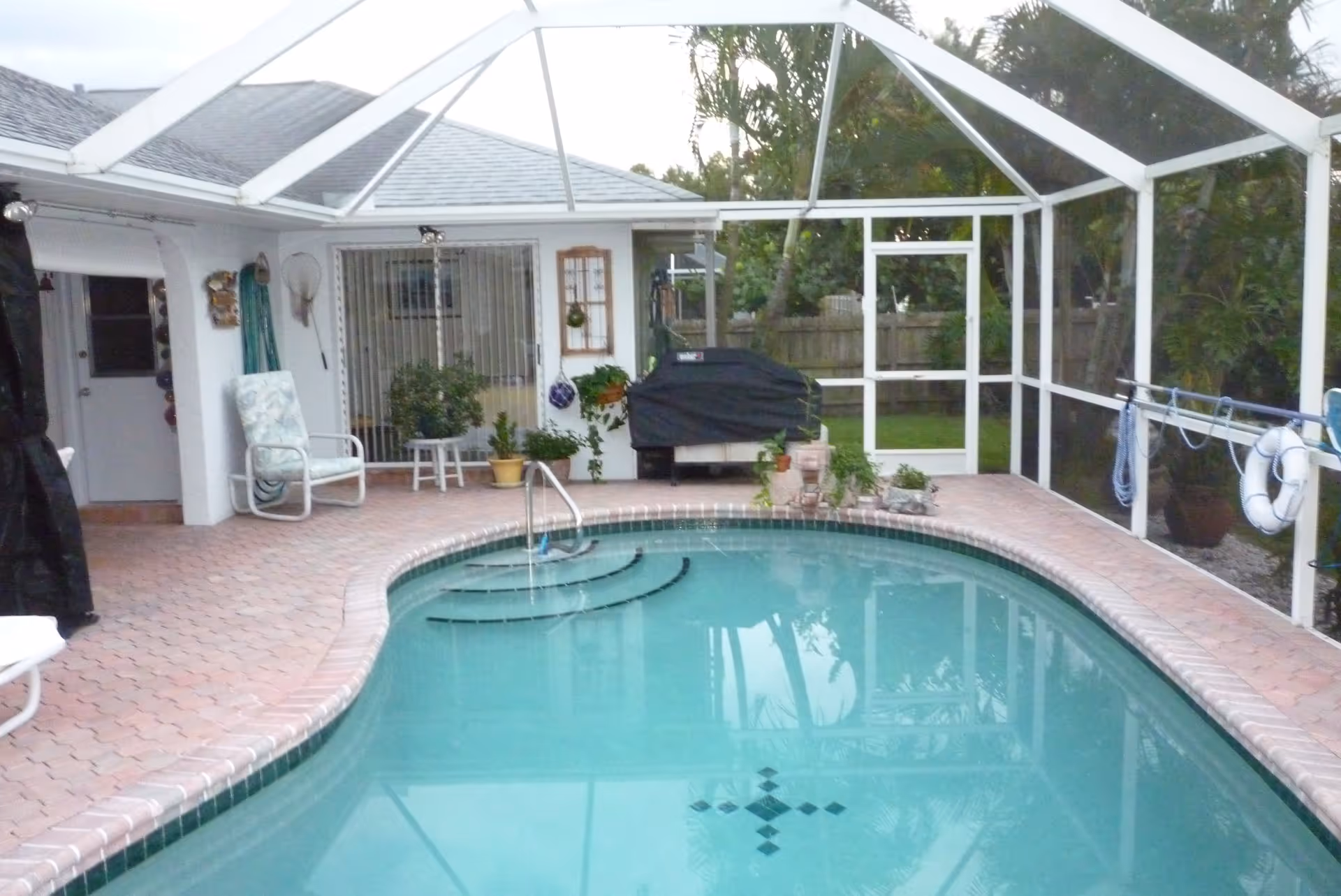 Screened-in backyard patio with a swimming pool, brick deck, lounge chairs, potted plants, and a covered grill.