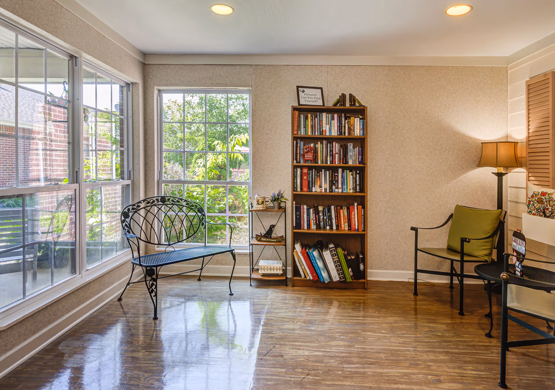 A cozy sitting area with large windows letting in natural light. The room features a black metal bench with decorative backrest, a wooden bookshelf filled with books, a small side table with decorative items, and a chair with a green cushion next to a floor lamp. The floor is polished wood, and the walls are light-colored.