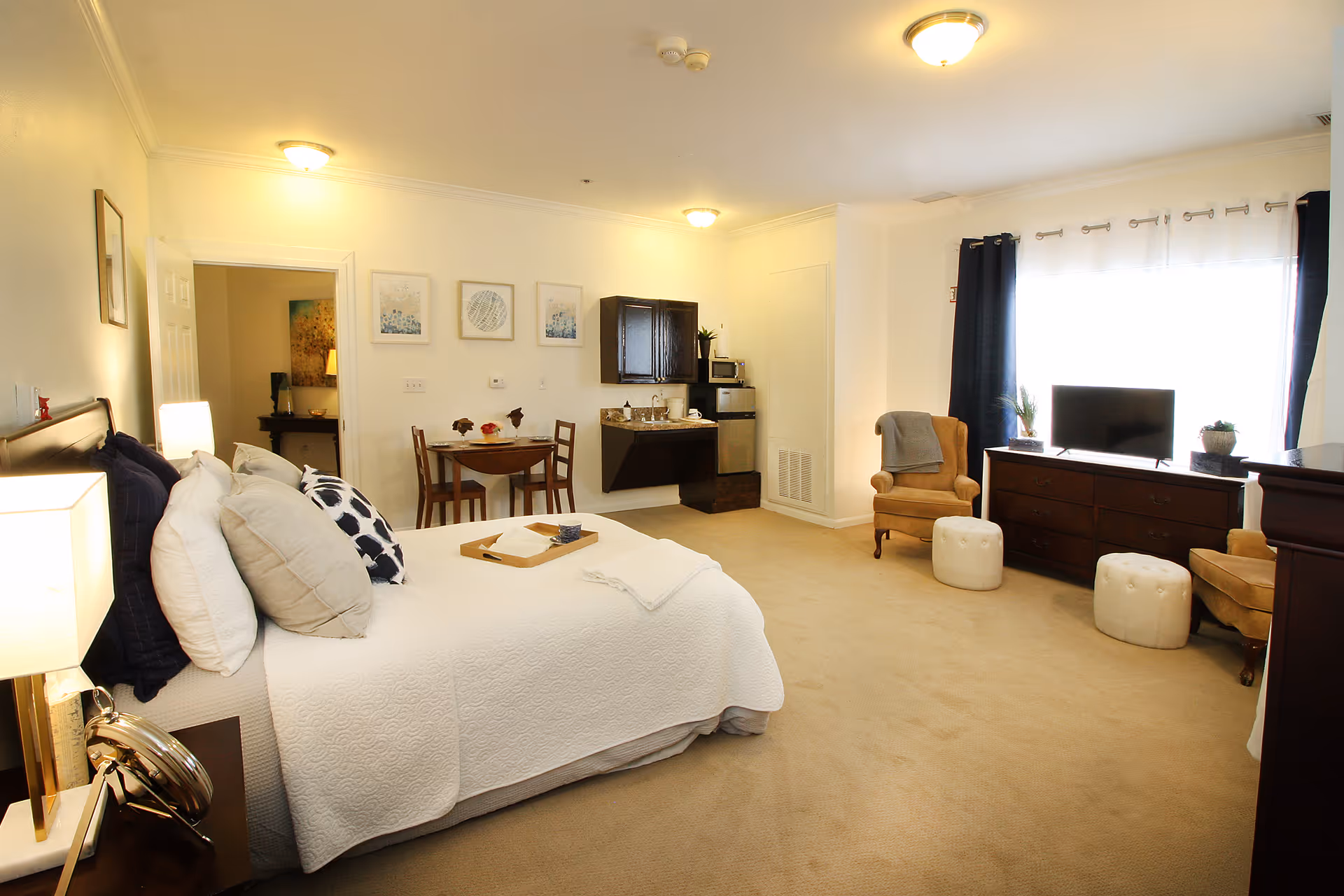 A spacious senior living bedroom featuring a large bed with multiple pillows and a white quilt, a small dining table with two chairs, a kitchenette with a microwave and mini fridge, two armchairs with ottomans, a dresser with a flat-screen TV, and large windows with dark curtains allowing natural light to fill the room.