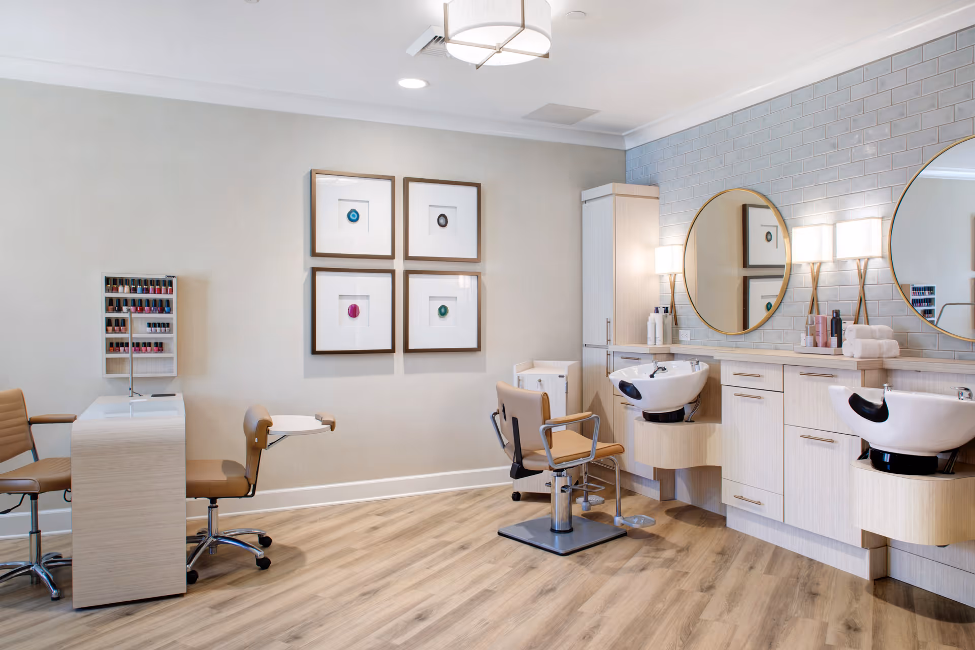 A bright and clean salon area in a senior living facility with two styling chairs in front of sinks and mirrors, a manicure station with nail polish display, light wood flooring, and modern decor including framed artwork and wall sconces.