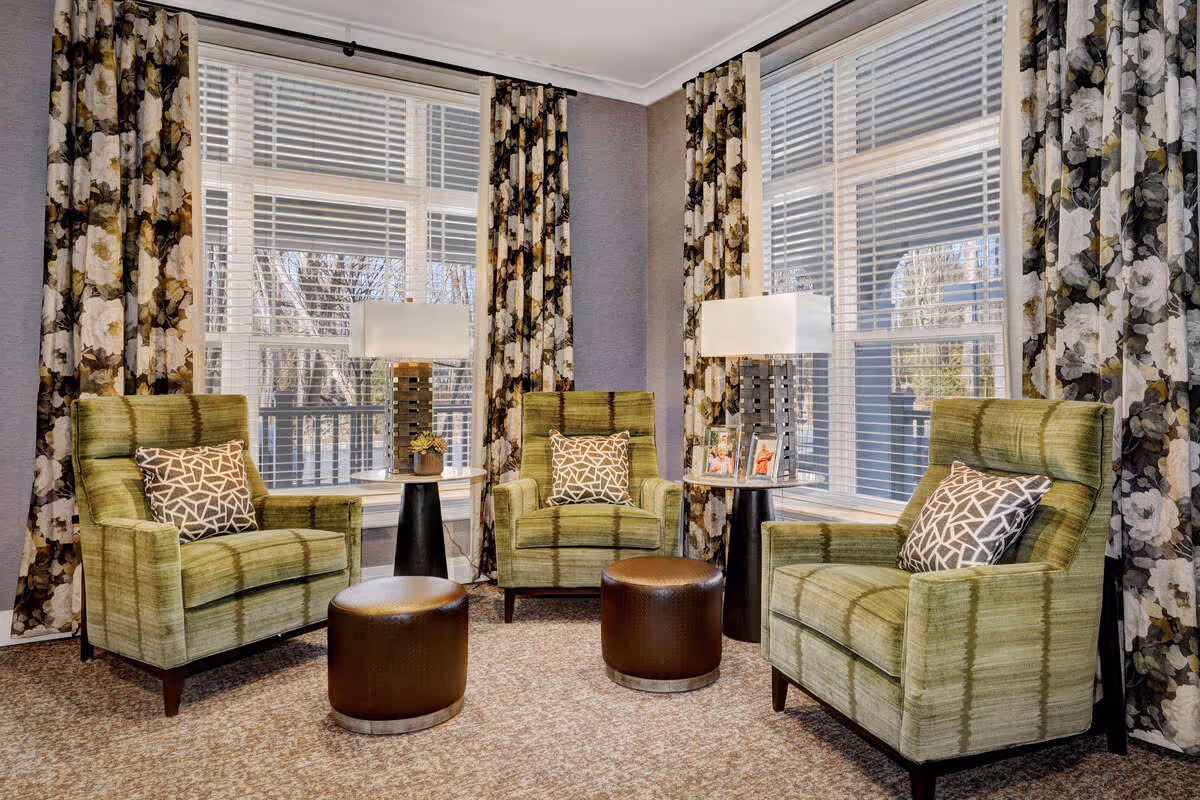 A cozy sitting area with three green upholstered armchairs, each with a patterned cushion, arranged around two small round brown ottomans. Behind the chairs are two side tables with modern lamps and framed photos. Large windows with white blinds and floral curtains allow natural light into the room.