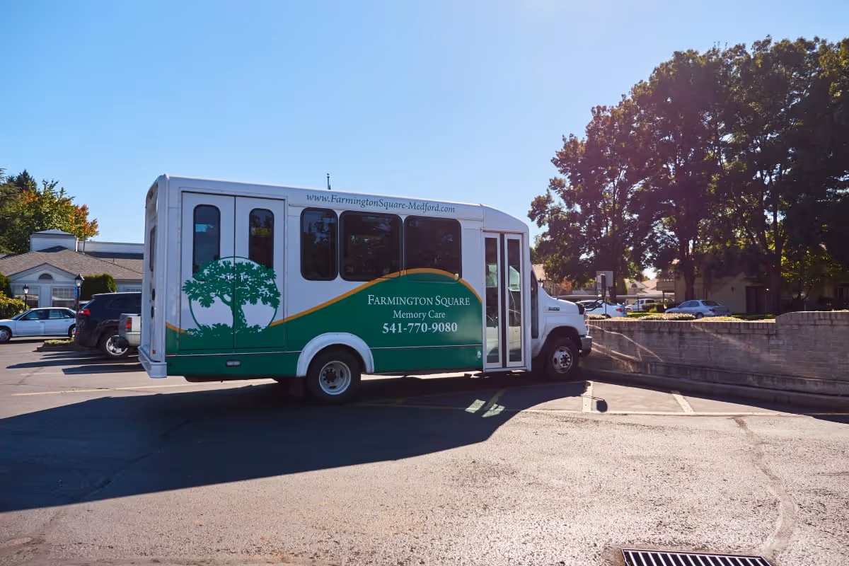 A white and green shuttle bus parked in a parking lot near a low brick wall with trees and other parked cars in the background. The bus has the text 'Farmington Square Memory Care 541-770-9080' and the website 'www.FarmingtonSquare-Medford.com' on its side.
