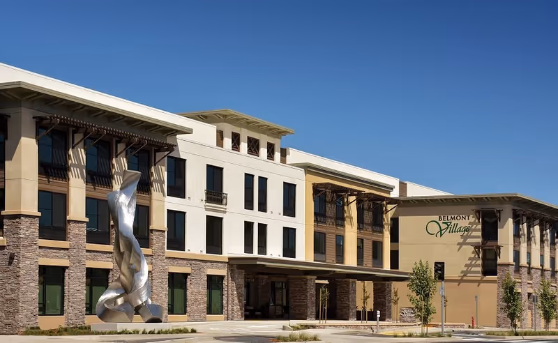 Exterior view of Belmont Village Senior Living Albany building with a modern architectural design, featuring stone and beige walls, multiple windows, a covered entrance, and a large abstract silver sculpture in front under a clear blue sky.