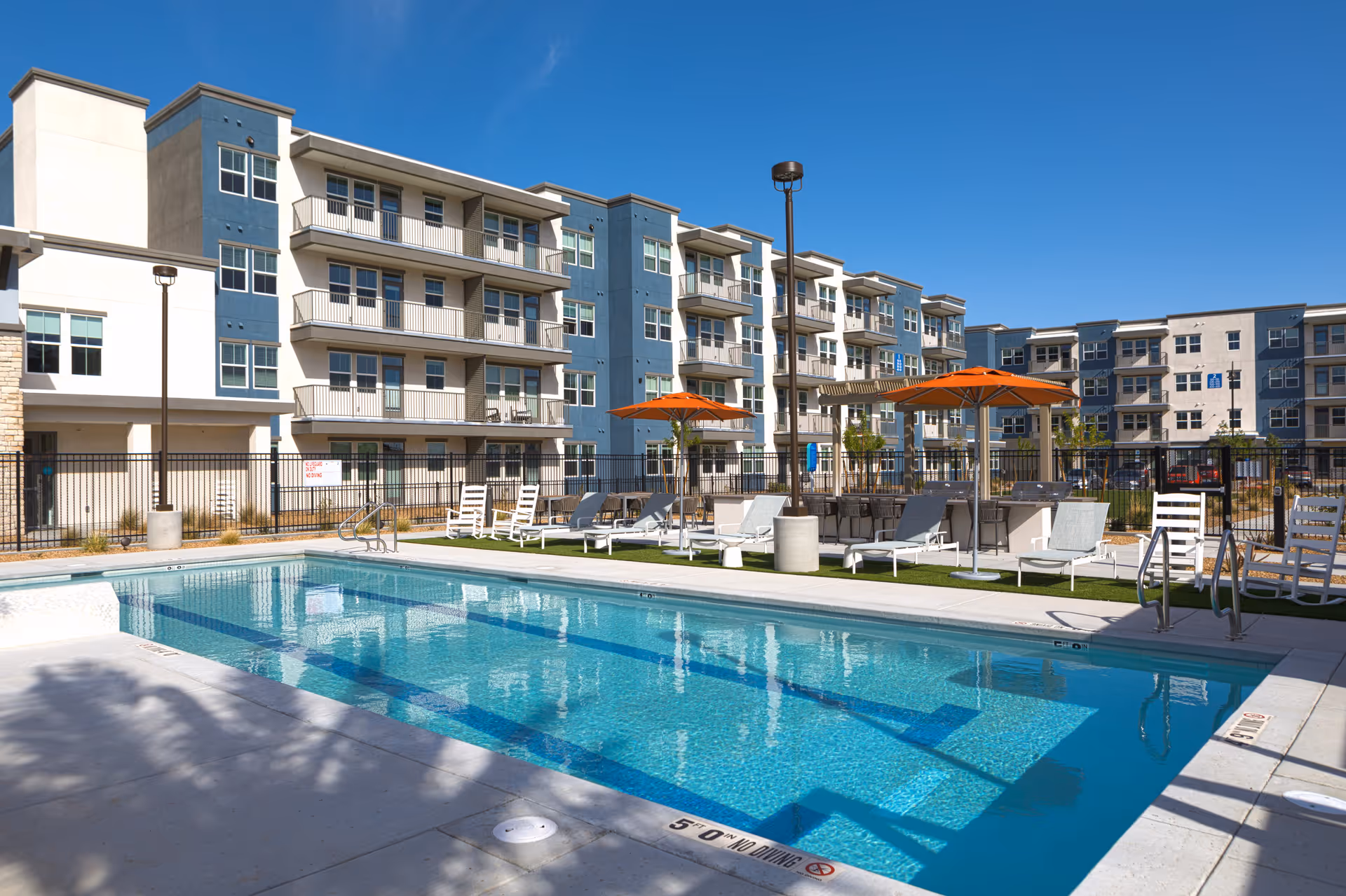 Outdoor swimming pool area with clear blue water, surrounded by white lounge chairs and orange umbrellas. In the background, there is a multi-story residential building with balconies under a clear blue sky.