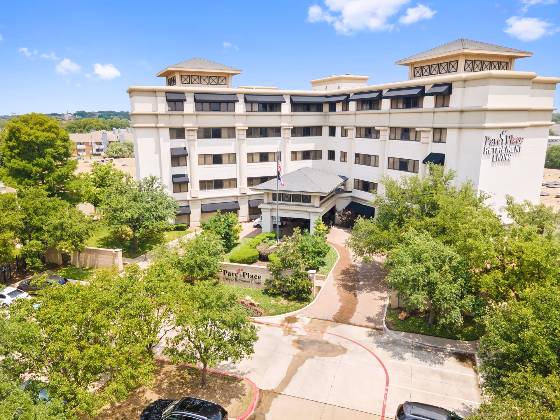 Exterior view of Parc Place Retirement Living, a multi-story senior living facility surrounded by trees and greenery under a blue sky with some clouds. The building has multiple windows with black awnings and a driveway leading to the entrance.