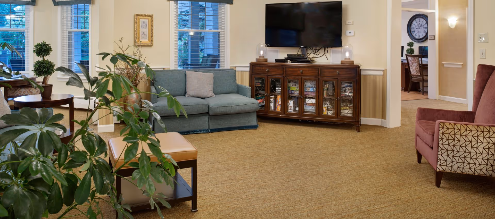 A cozy living room area with a blue loveseat, a wooden cabinet with glass doors holding books and games, a flat-screen TV mounted on the wall above the cabinet, a maroon armchair with patterned sides, and several green plants. There are windows with white blinds letting in natural light and a glimpse into an adjacent room with a large wall clock and a desk with a chair.