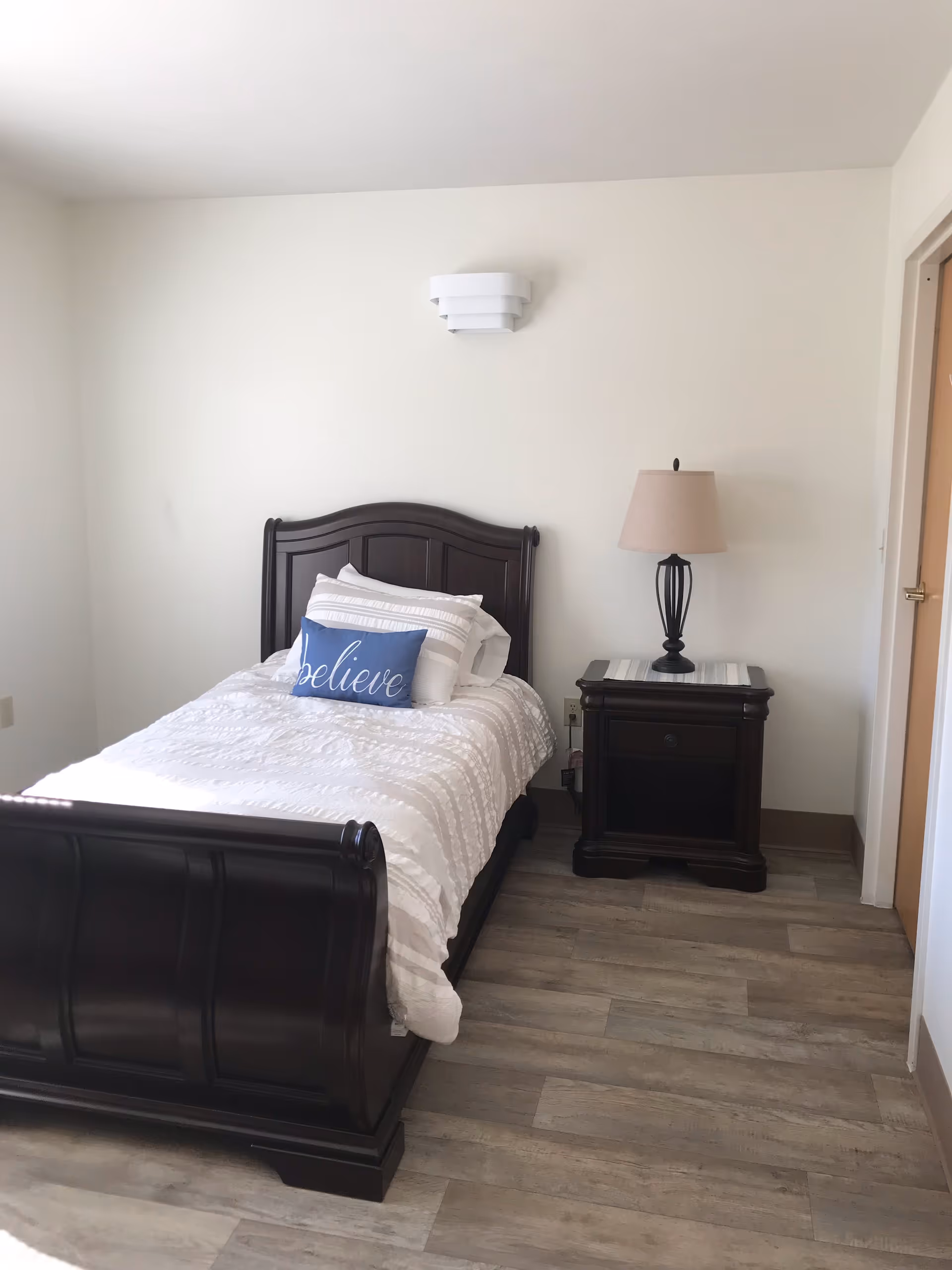 A bedroom with a dark wooden bed frame and white bedding, including a blue pillow with the word 'believe' on it. Next to the bed is a matching dark wooden nightstand with a lamp on top. The room has light-colored walls and wood-look flooring.