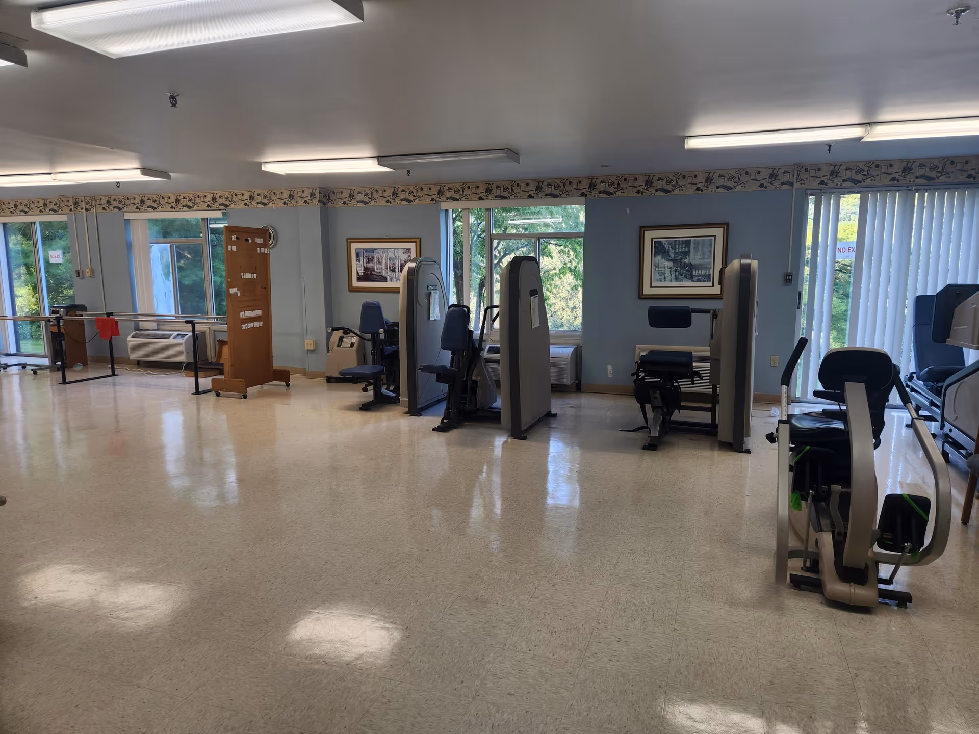 A spacious exercise room with various fitness equipment including stationary bikes and strength training machines. The room has large windows letting in natural light, framed pictures on the walls, and a polished floor. There is also a wooden board with instructions or information near the windows.