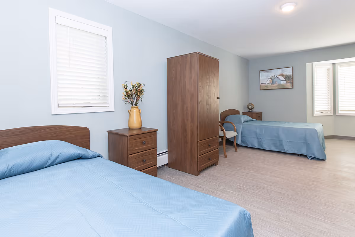 Spacious twin bedroom with two beds covered in blue bedspreads, a wooden nightstand and wardrobe, a chair, framed art, and windows with blinds.