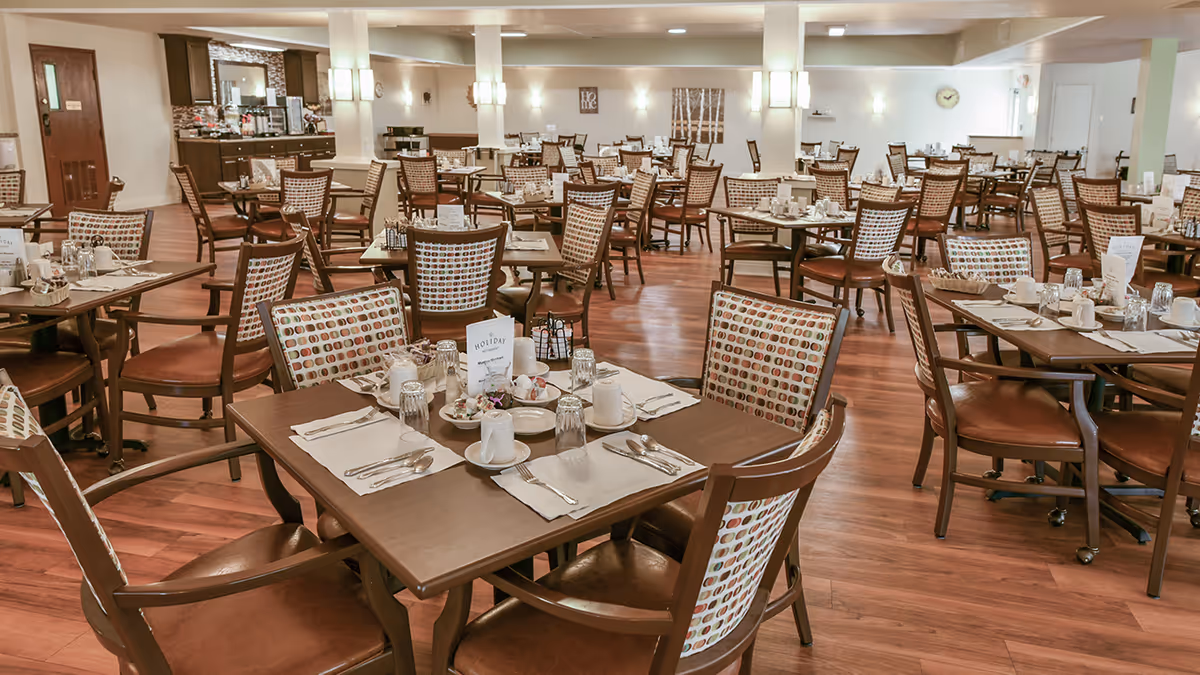 A spacious dining room with multiple wooden tables and chairs arranged neatly. Each table is set with placemats, utensils, cups, glasses, and small condiment holders. The chairs have patterned backs and leather seats. The room has wooden flooring, soft lighting, and neutral-colored walls with minimal decorations.