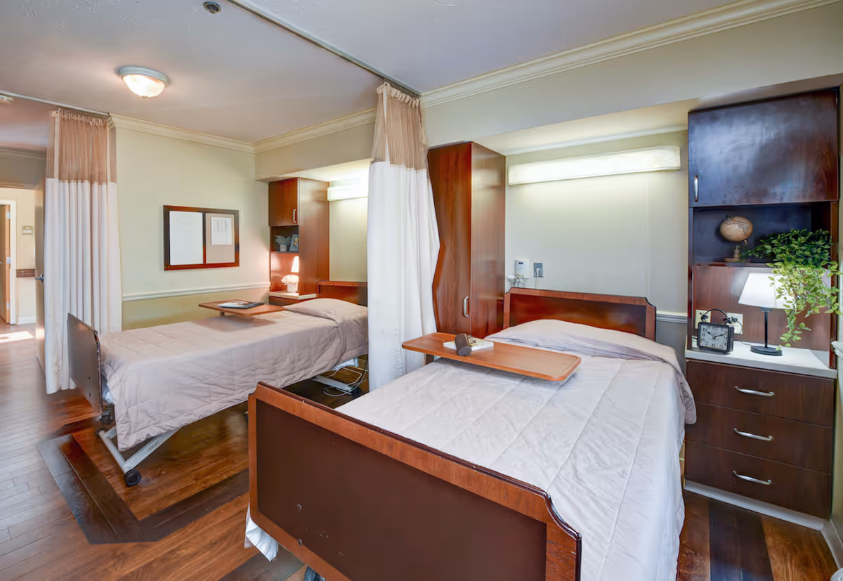 A shared bedroom in a senior living facility with two hospital-style beds separated by a curtain. Each bed has a wooden headboard and footboard, beige bedding, and an overbed table. There are built-in wooden cabinets and shelves with a lamp, clock, and decorative plant. The room has wooden flooring and soft lighting.