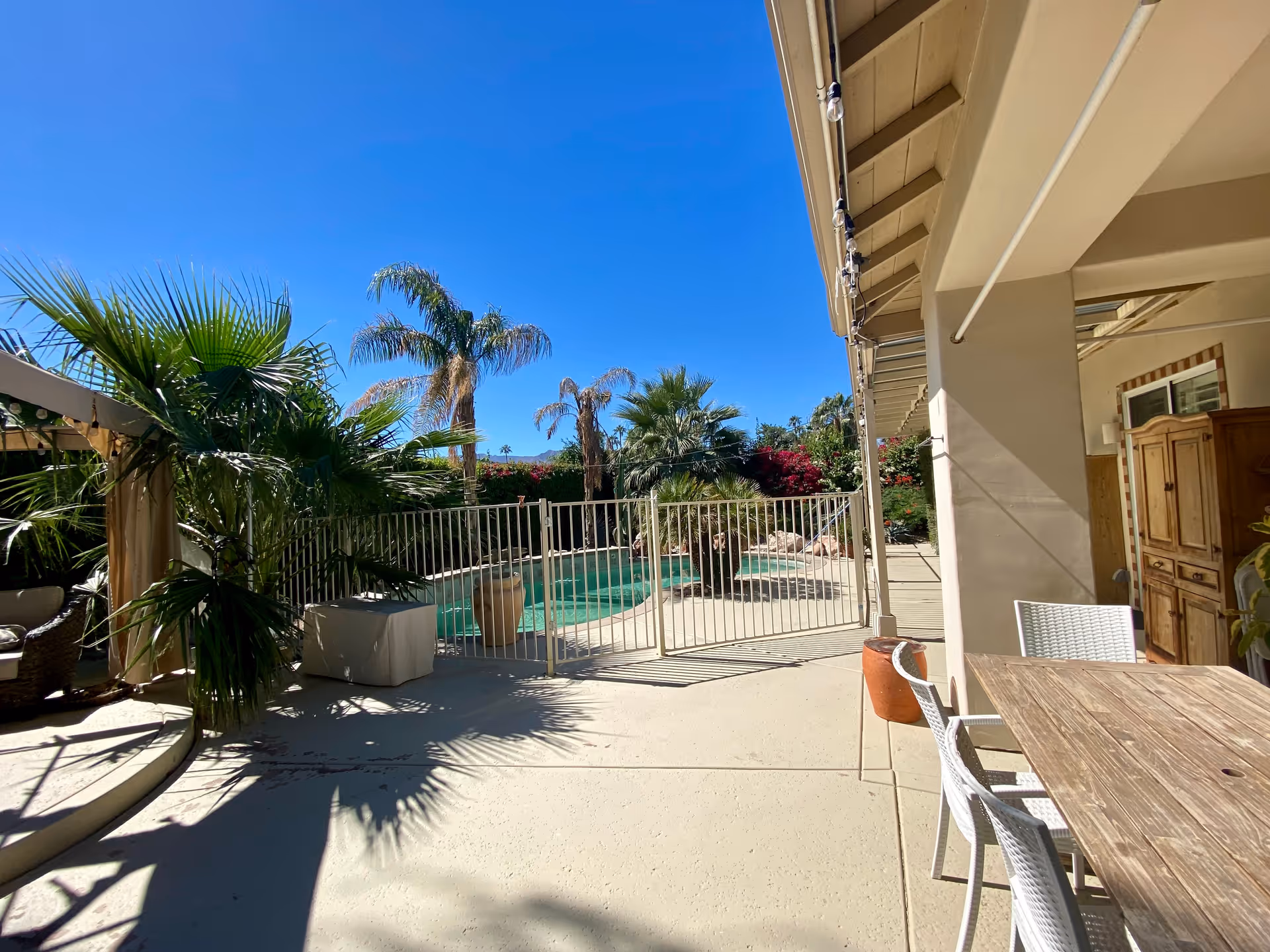 Outdoor patio area with a wooden table and white chairs under a covered section. Beyond the patio is a fenced swimming pool surrounded by palm trees and other greenery under a clear blue sky.
