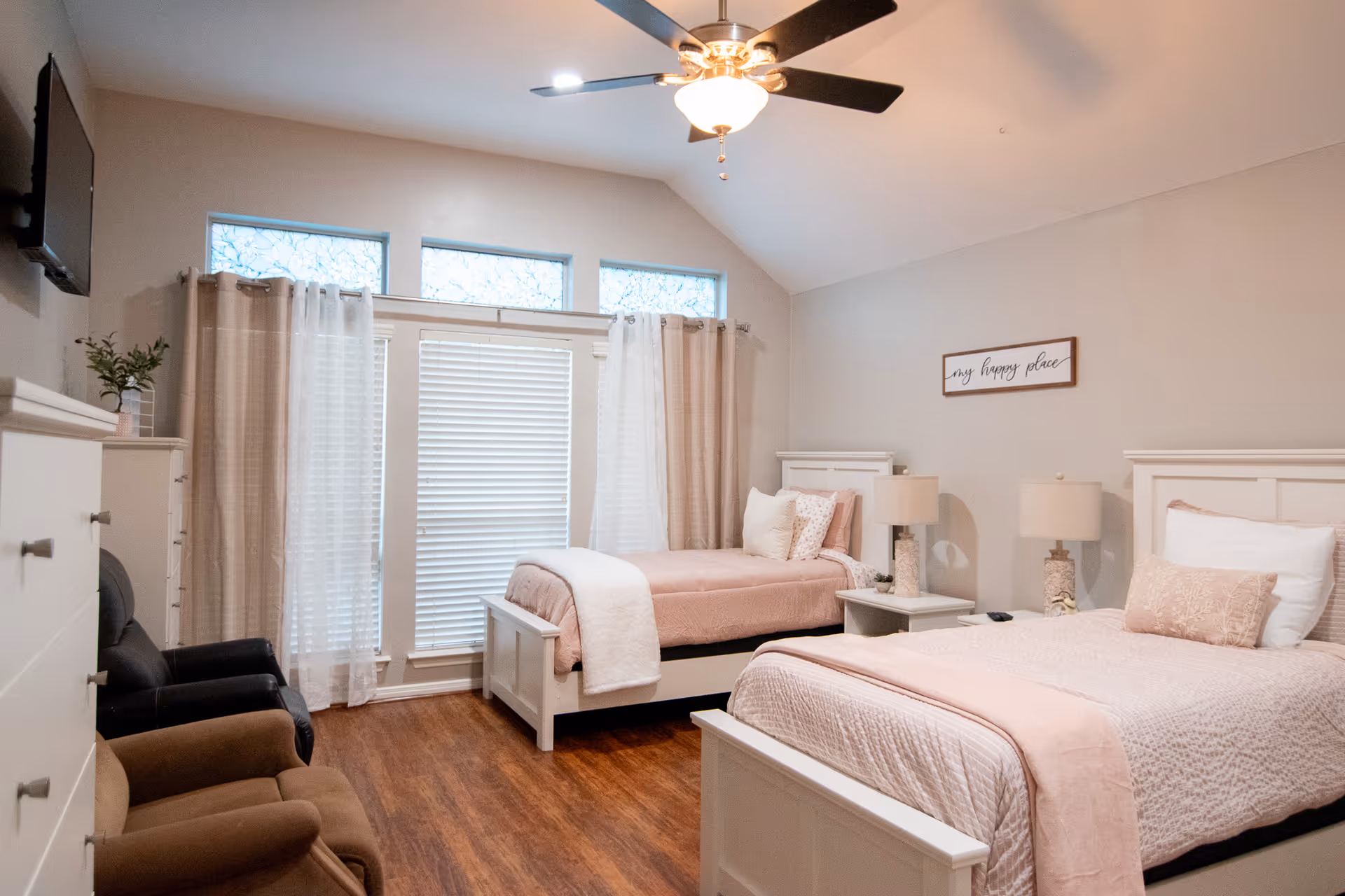A bright and cozy bedroom with two twin beds featuring light pink bedding and white headboards. Between the beds is a nightstand with two matching lamps. The room has wood flooring, a ceiling fan with lights, a wall-mounted TV, and large windows with blinds and curtains. There are two armchairs and a dresser on the left side of the room. A framed sign on the wall reads 'my happy place'.