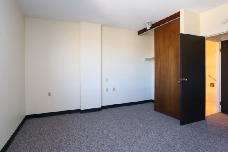 Empty room with beige walls, gray carpet, a wooden closet door partially open, and an open doorway leading to a bathroom with a bathtub visible.