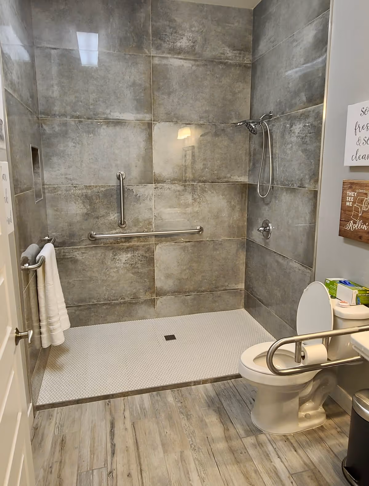 Accessible bathroom with a walk-in shower featuring large gray tiles, a handheld showerhead, and grab bars. There is a toilet with a support rail, a towel rack with white towels, and decorative wall signs. The floor has wood-like tiles.