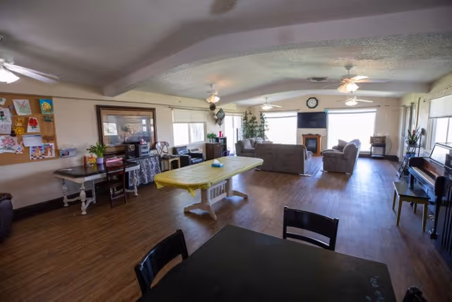 A spacious common area in a senior living facility with wooden flooring, several chairs and tables, a piano on the right side, a bulletin board with artwork on the left wall, and a seating area with sofas and a TV near large windows at the far end.