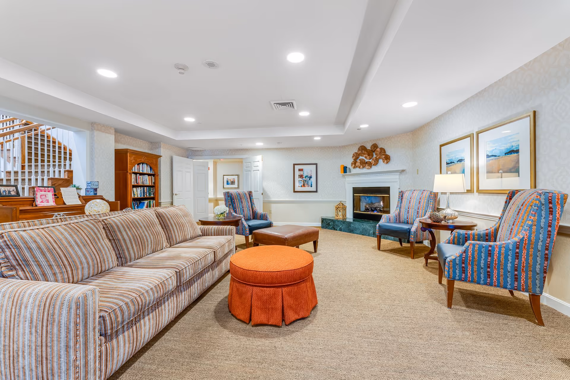 A bright and spacious living room area with a striped sofa, an orange round ottoman, and three patterned armchairs arranged around a fireplace. The room features beige carpet, framed artwork on the walls, a wooden bookshelf, and a staircase in the background.