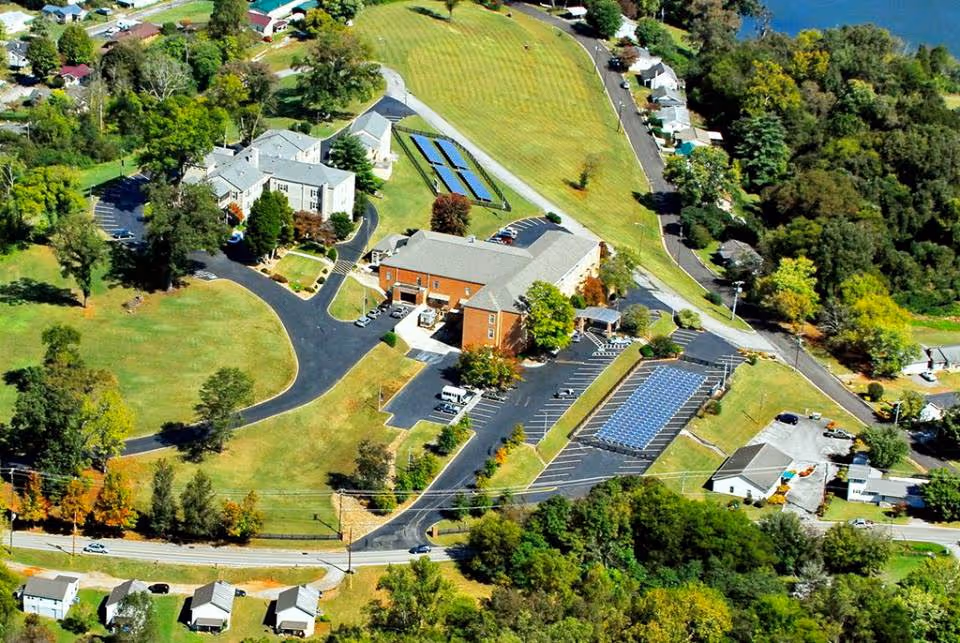Aerial view of Lakewood Place senior living facility showing multiple buildings, parking lots with solar panels, surrounding greenery, and nearby houses along a road.