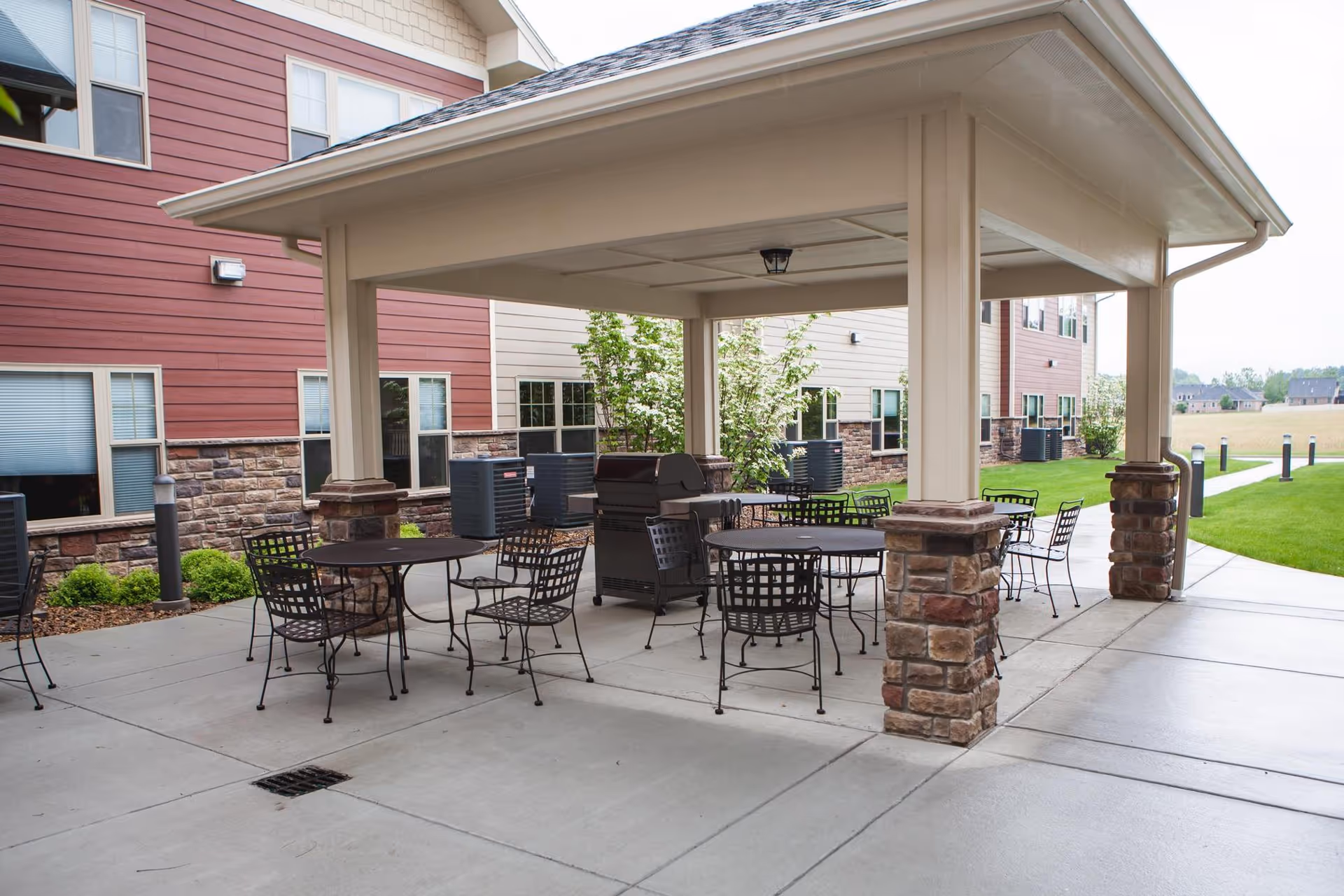Outdoor covered patio area with several round metal tables and chairs, a barbecue grill, and a walkway leading to a grassy area. The patio is adjacent to a building with red and beige siding and stone accents.