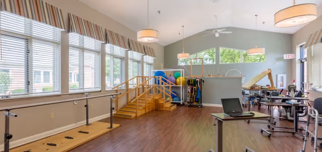 A spacious rehabilitation or physical therapy room with wooden flooring, large windows with blinds, parallel bars for walking exercises, a small wooden staircase, exercise balls, and various therapy equipment. The room is well-lit with ceiling lights and has a high ceiling with a fan.