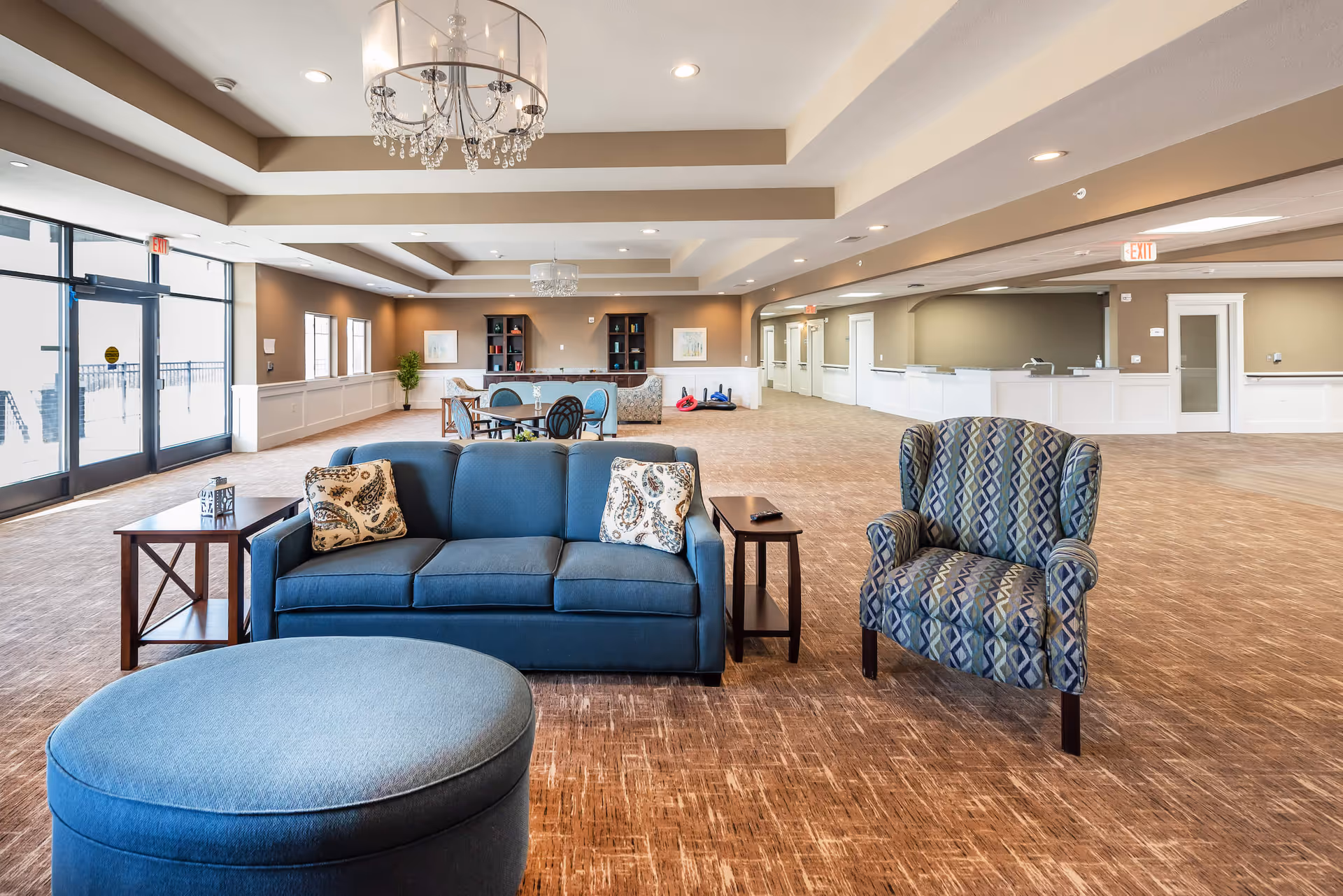 A spacious and well-lit senior living facility common area with a blue sofa, patterned armchair, and round ottoman in the foreground. There are side tables next to the sofa, a dining table with chairs in the background, and bookshelves along the far wall. The room features large windows, chandeliers, and a beige and white color scheme with carpeted flooring.