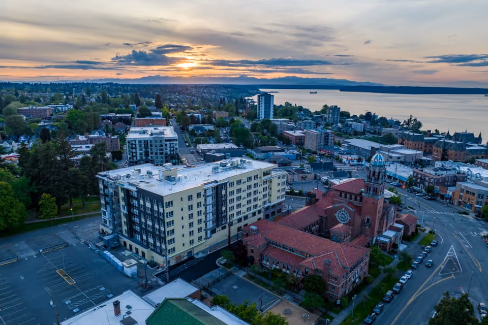 Aerial view of a cityscape at sunset featuring a large multi-story residential building next to a historic church with a red-tiled roof and a distinctive dome. The scene includes surrounding streets, parked cars, trees, and a body of water in the background under a partly cloudy sky.