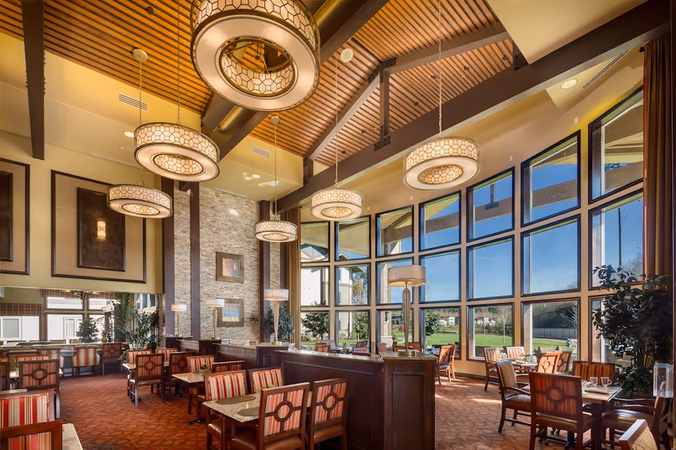 Spacious dining room with a high wooden beamed ceiling, large circular pendant lights, floor-to-ceiling windows and multiple tables with striped chairs overlooking a lawn.
