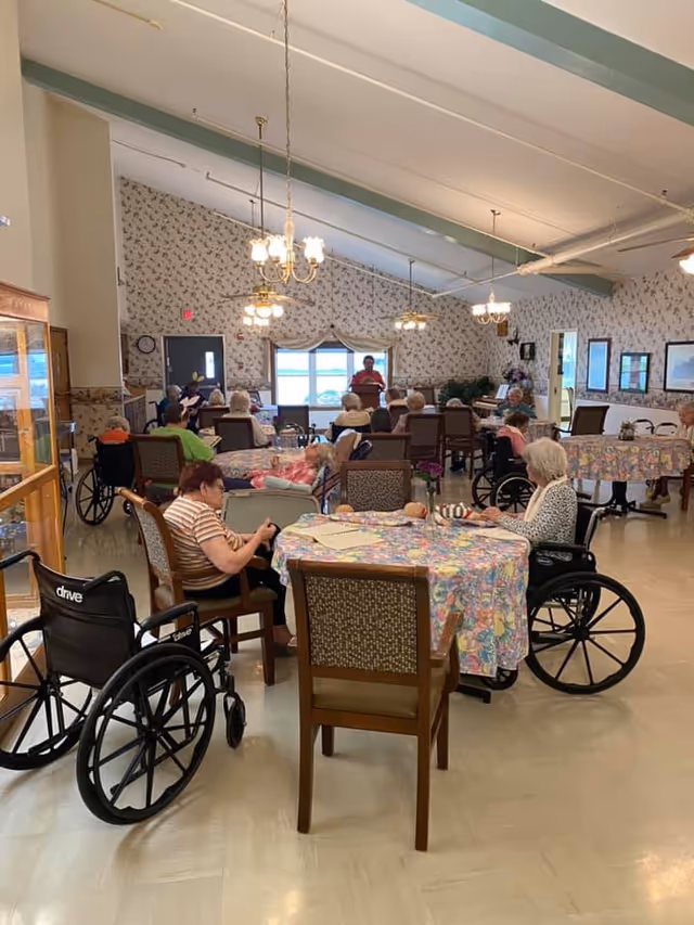 A group of elderly people, some in wheelchairs, seated around tables covered with floral tablecloths in a large, well-lit room with floral wallpaper and chandeliers. A person stands at the front of the room near a window, possibly addressing the group. The room has a cozy, communal atmosphere.