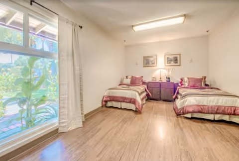 A bright bedroom with two twin beds featuring matching beige and purple bedding. Between the beds is a dark wooden nightstand with a lamp and decorative items. Two framed pictures hang on the wall above the nightstand. A large window with white curtains allows natural light to fill the room and offers a view of greenery outside. The floor is a light wood laminate.
