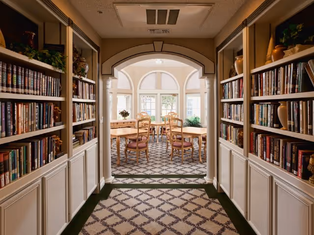 A hallway lined with bookshelves opens through an arched doorway into a sunlit dining area with wooden tables and chairs.