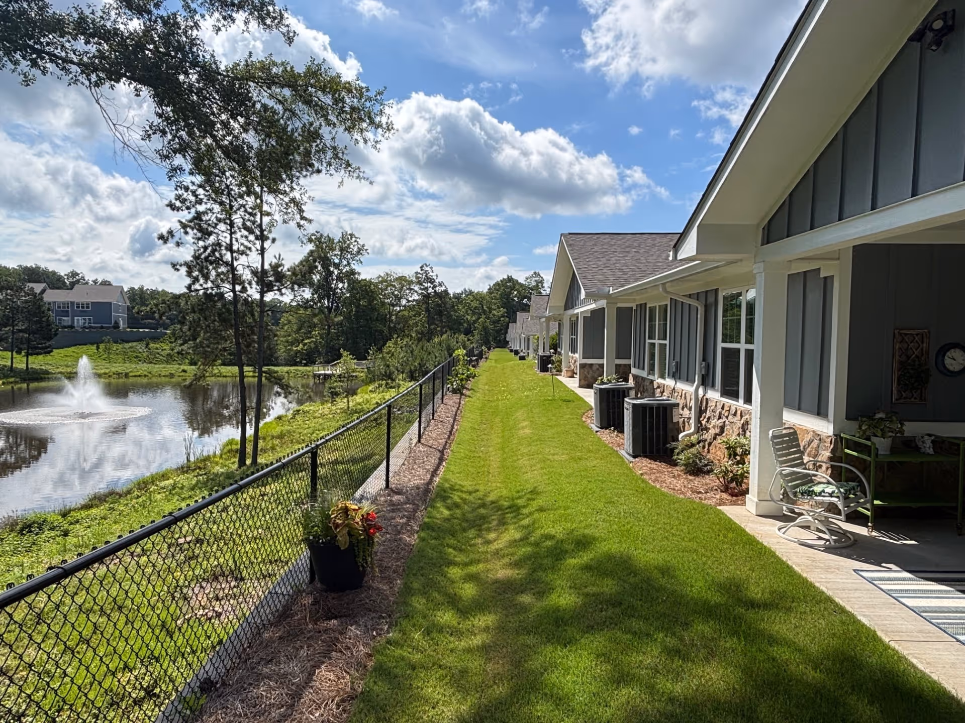 Row of single-story assisted-living units with porches and a fenced grassy walkway overlooking a pond with a fountain under a partly cloudy sky.