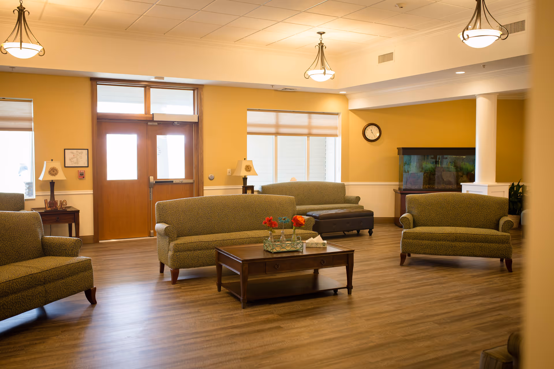 A spacious and well-lit living room area with multiple green upholstered sofas arranged around a wooden coffee table with decorative flowers. The room has wooden flooring, yellow walls, ceiling lights, a clock on the wall, and a large fish tank in the corner. There are windows and a wooden door with glass panels letting in natural light.