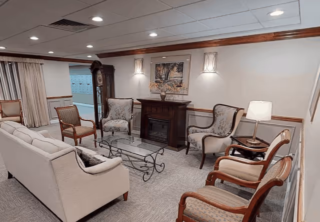 A cozy assisted living facility common area with a beige sofa, several upholstered armchairs, a glass coffee table with a decorative metal frame, a wooden electric fireplace, and a table lamp. The room has carpeted flooring, recessed ceiling lights, wall sconces, and a framed painting above the fireplace.