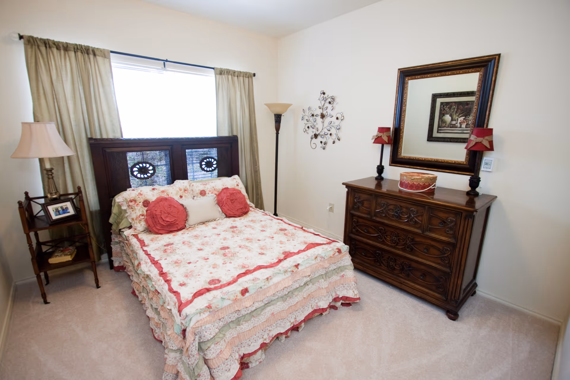 A cozy bedroom with a floral-patterned bedspread and two round decorative pillows on a wooden bed frame. There is a wooden nightstand with a lamp and a framed photo on the left side of the bed. A tall floor lamp stands in the corner next to a wall decorated with a metal wall art piece. On the right side, there is a wooden dresser with intricate carvings, topped with two small lamps and a decorative box, and a large framed mirror hanging above it. The room has beige carpet and light-colored walls with a window covered by curtains behind the bed.