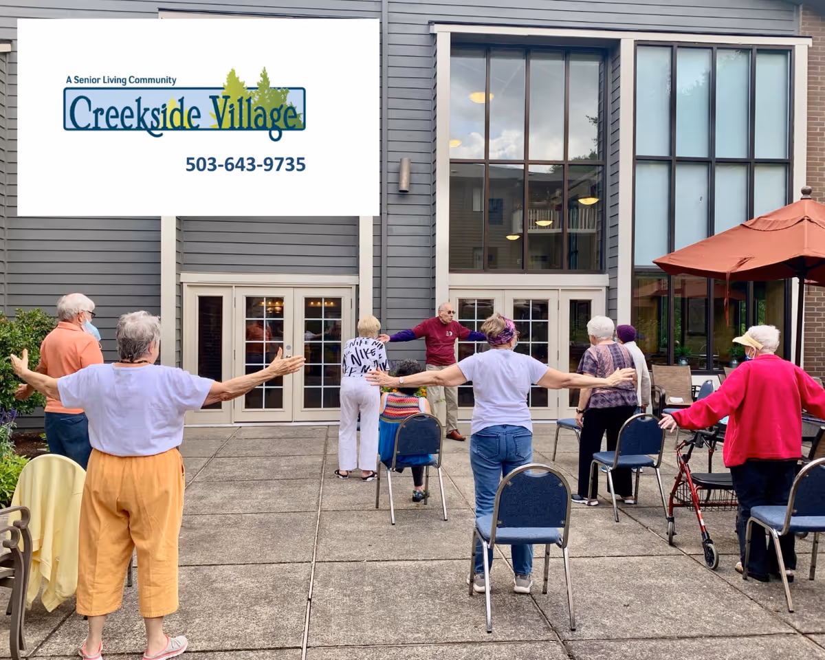 A group of elderly people participating in an outdoor exercise session on a patio area in front of a building with large windows. Some participants are standing with arms outstretched, while others are seated on chairs. The setting appears to be a senior living community.
