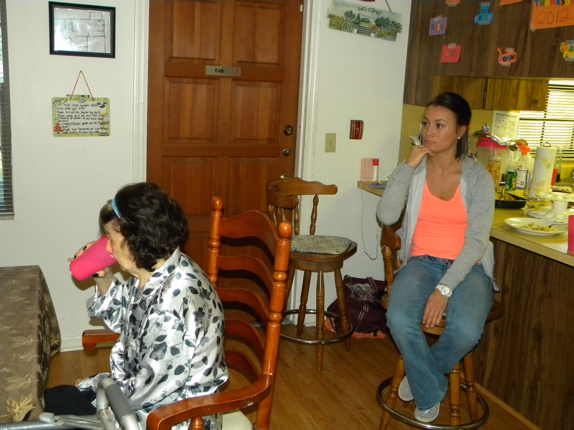 An elderly woman drinking from a pink cup seated at a dining table, and a younger woman sitting on a wooden bar stool near a kitchen counter in a room with wooden floors and a wooden door marked 'Exit'. The kitchen counter has various items including plates, cups, and a soda can. The walls have decorations and signs, including a 'Welcome' sign and colorful paper notes.